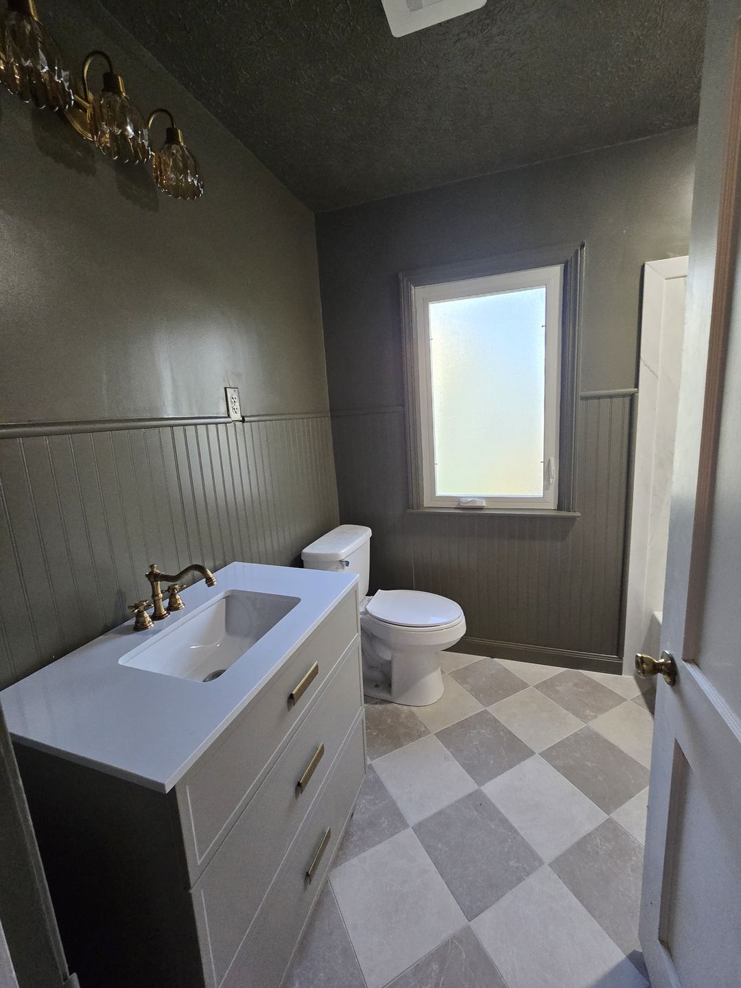 Finished bathroom with olive green walls and beadboard, white trim around the window, and a checkerboard floor