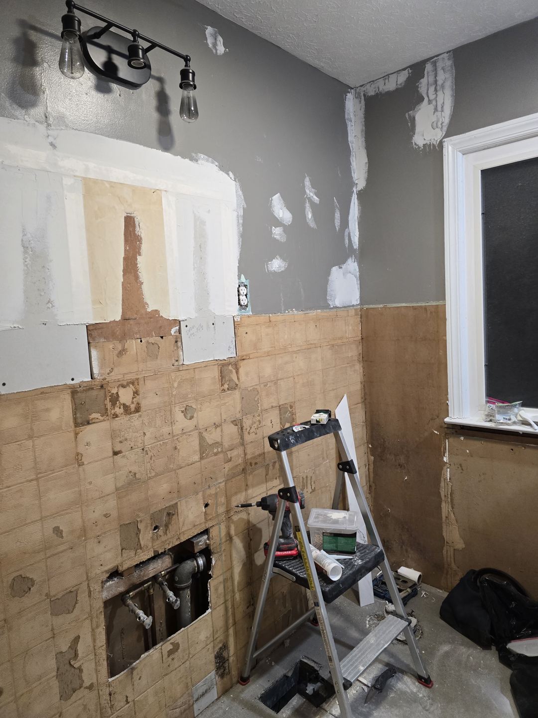 Bathroom wall with the old tile torn off, bare patches of drywall and a stepladder in the middle of the room
