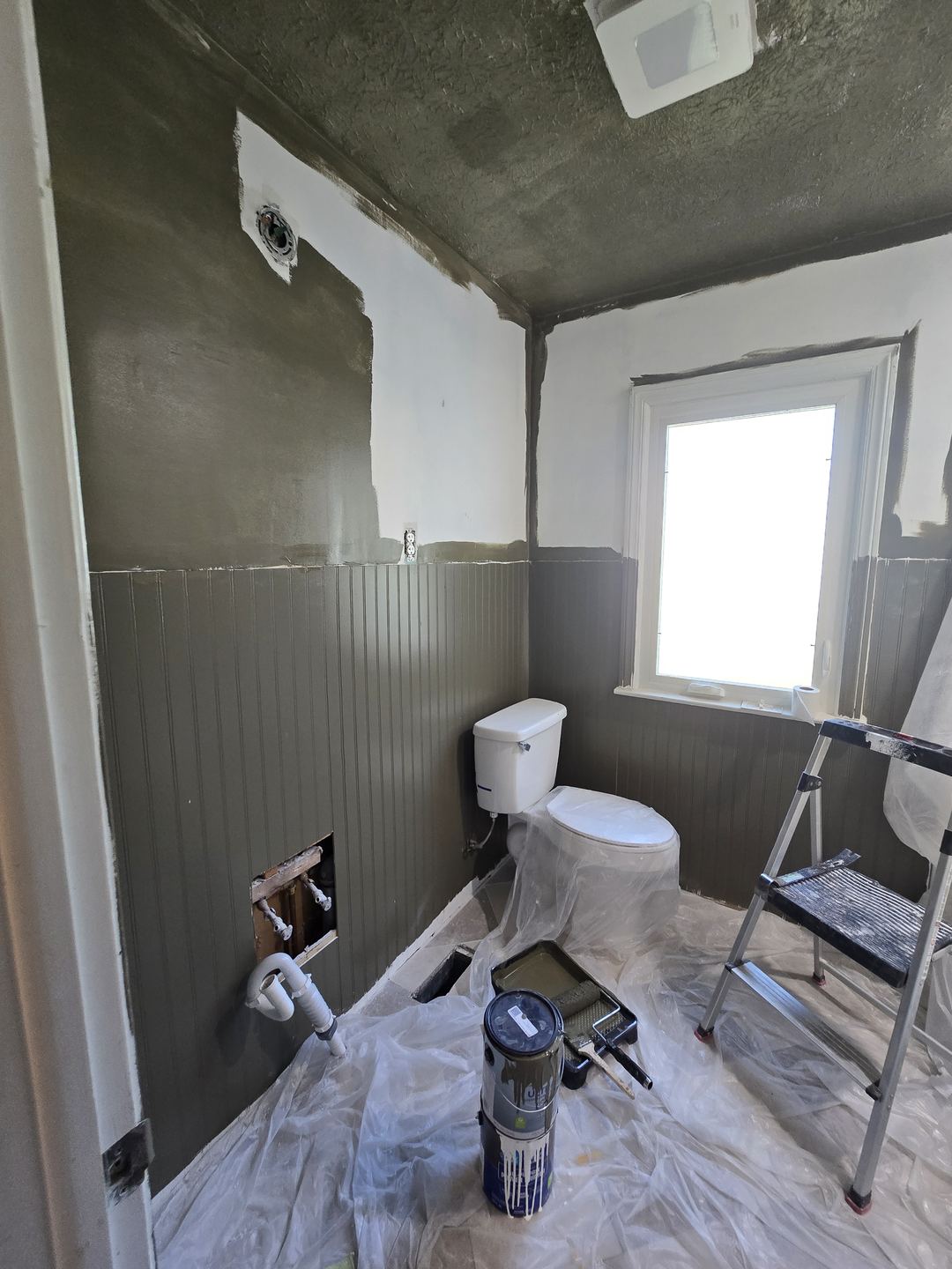 Bathroom mid-paint with plastic covering the floor and toilet, a paint can and roller tray on the floor